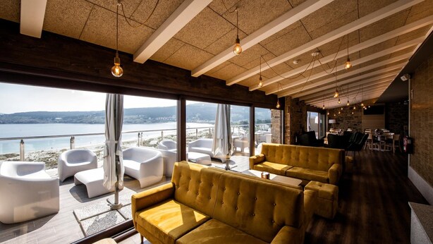 A modern interior with a large window overlooking a beach, featuring a yellow sofa and white chairs. - Apartamentos Vida Mar de Laxe (Laxe)