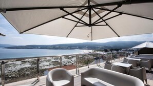 A terrace with white chairs and tables, a large umbrella, and a view of the sea and distant hills. - Apartamentos Vida Mar de Laxe (Laxe)