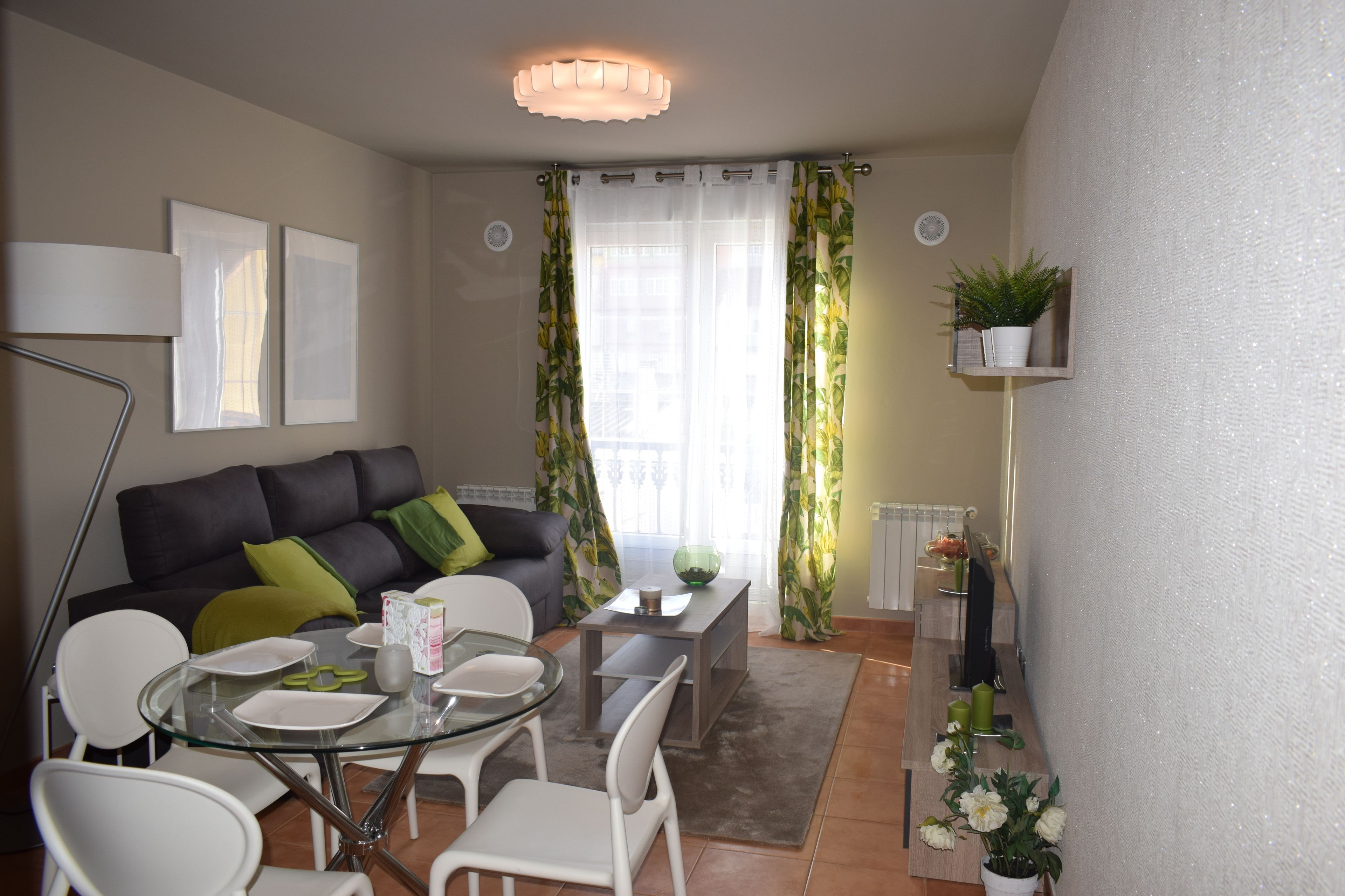 A modern living room with a sofa, dining table, chairs, and a coffee table. There is a window with curtains, a floor lamp, and a small shelf with a plant.