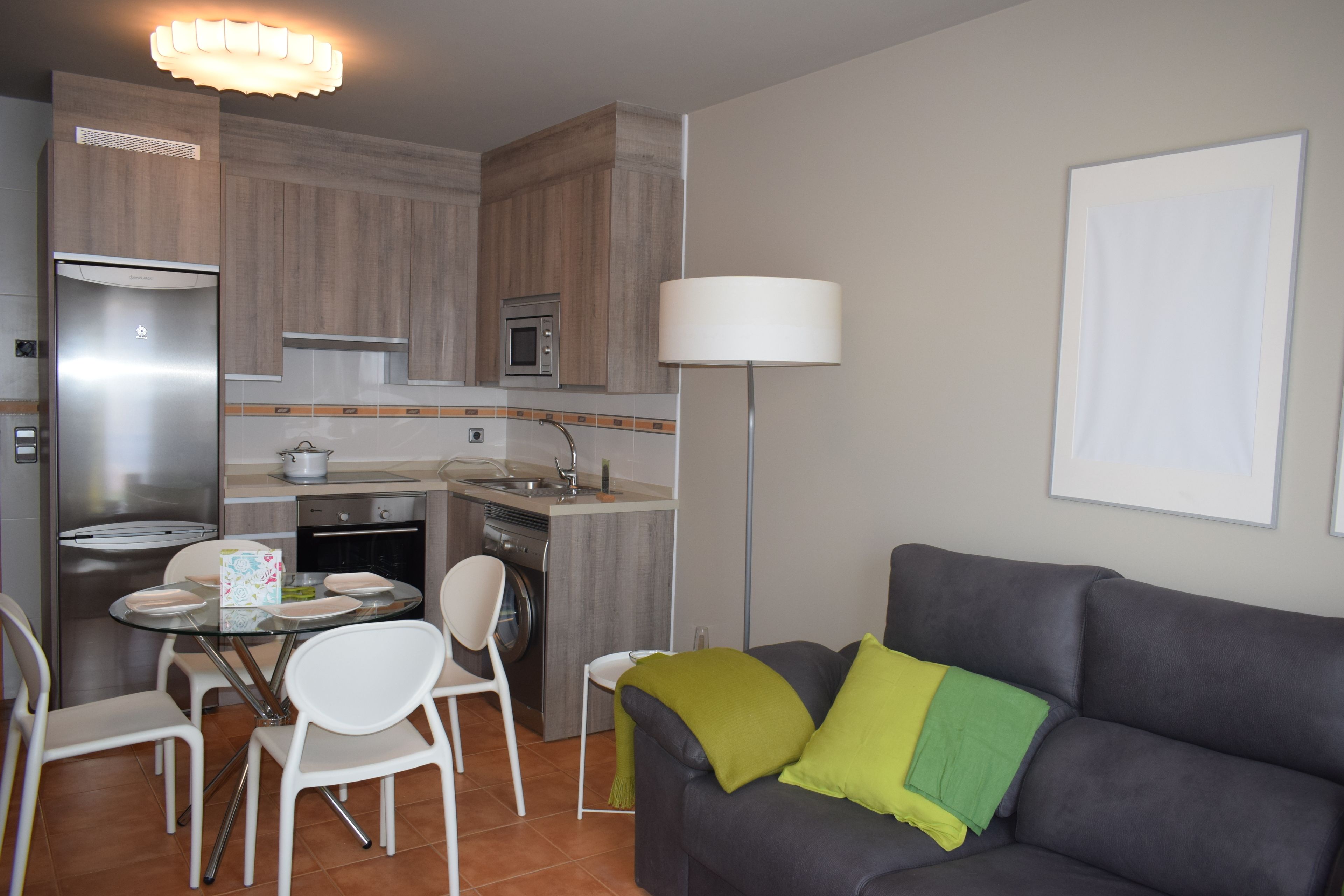Un espacio habitable compacto con kitchenette, una zona de comedor con sillas, un sofá con cojines y una lámpara de pie.