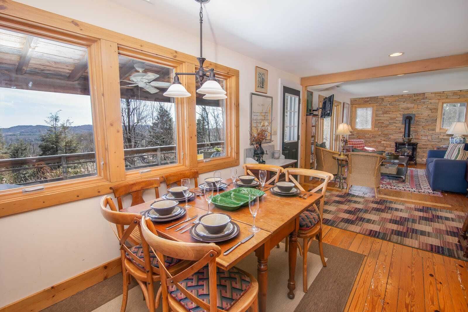 A dining area with a wooden table set for four, large windows, and a stone wall in the background.