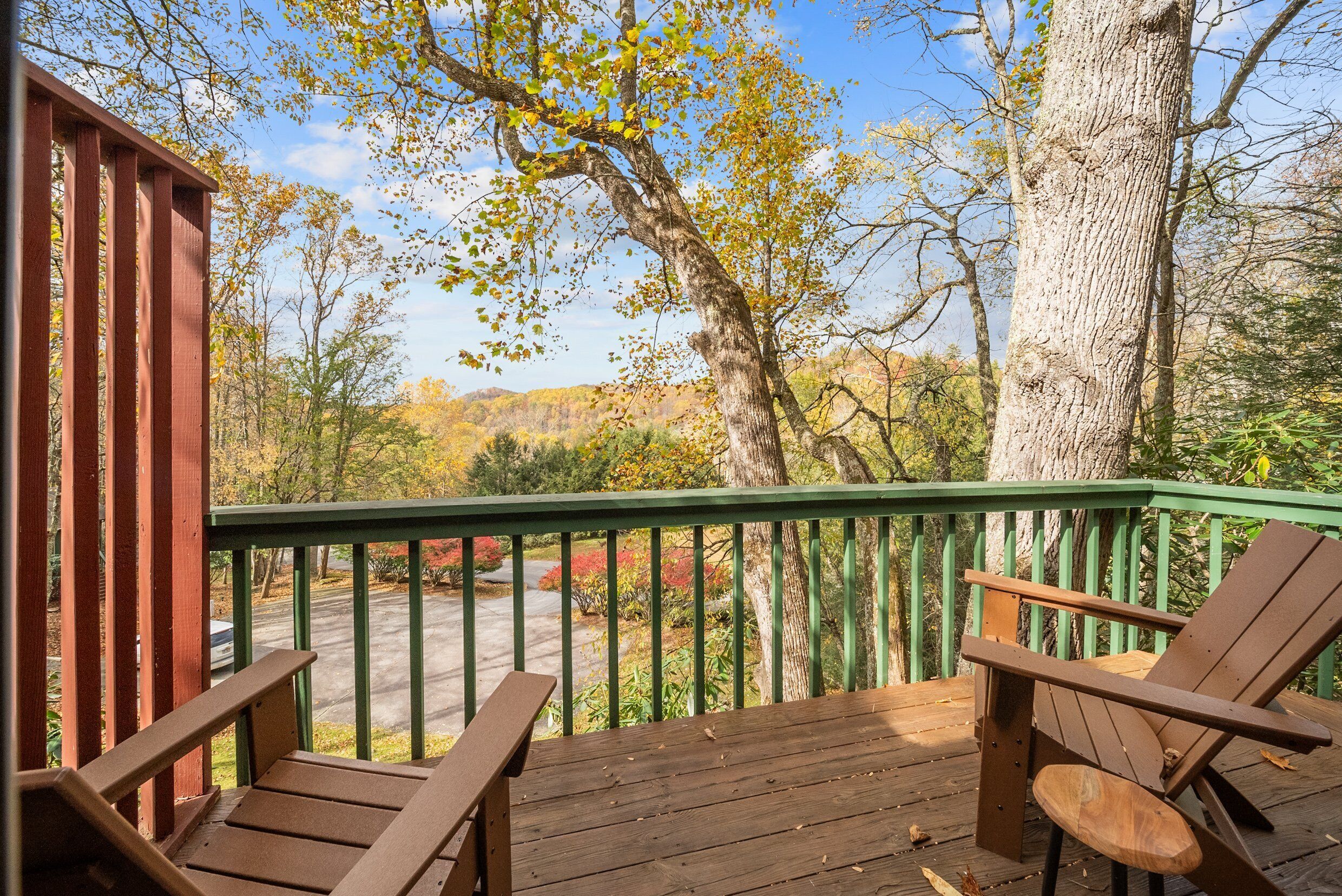 A wooden deck with two Adirondack chairs and a green railing overlooking a scenic landscape with trees and a road.
