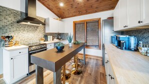 A modern kitchen with a wooden ceiling, a central island with stools, white cabinets, and a large window with blinds. - Vacation Cottage in Yonahlossee Resort (Boone)