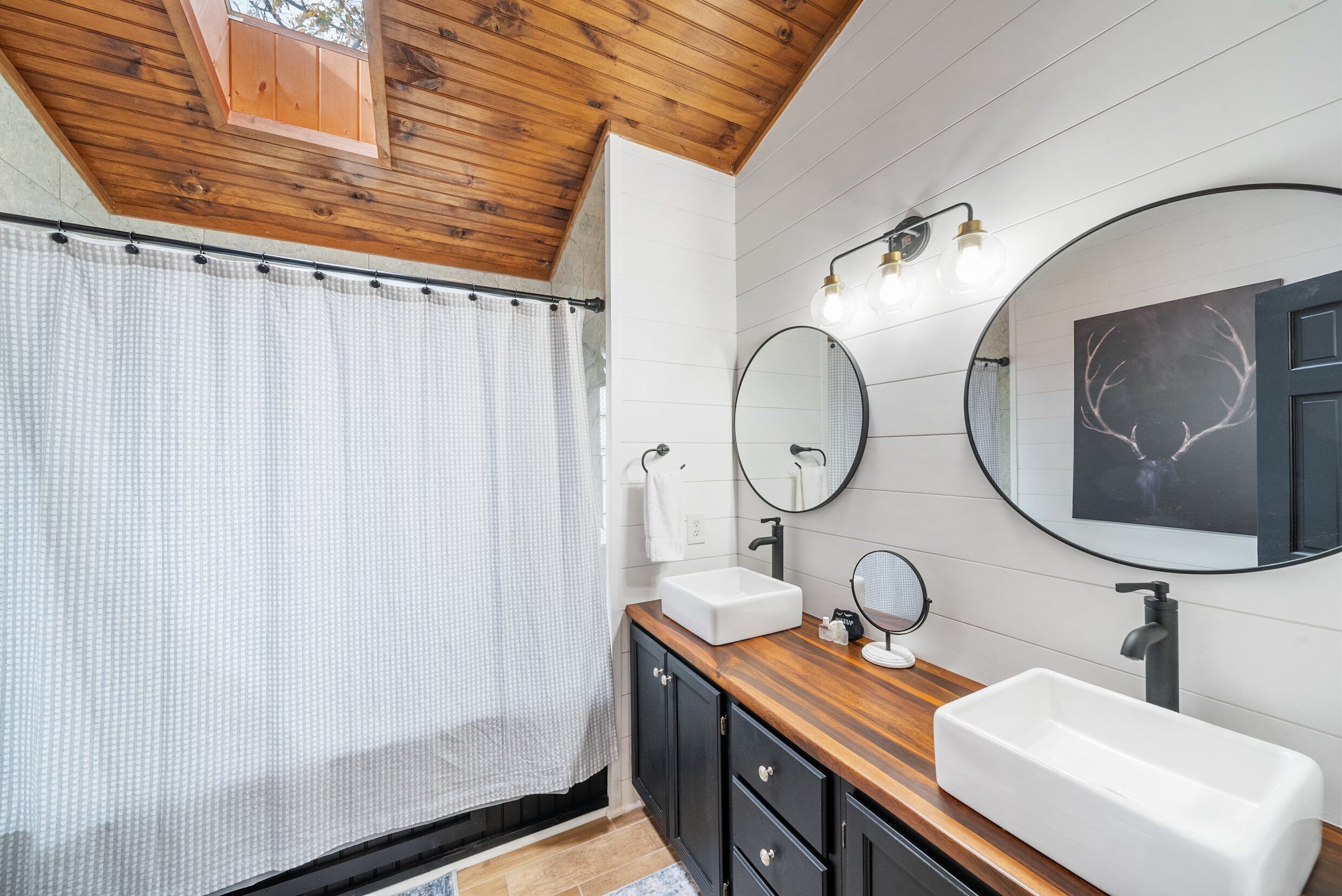 Une salle de bain avec un plafond en bois, des murs recouverts de carreaux blancs, un grand miroir et deux lavabos.