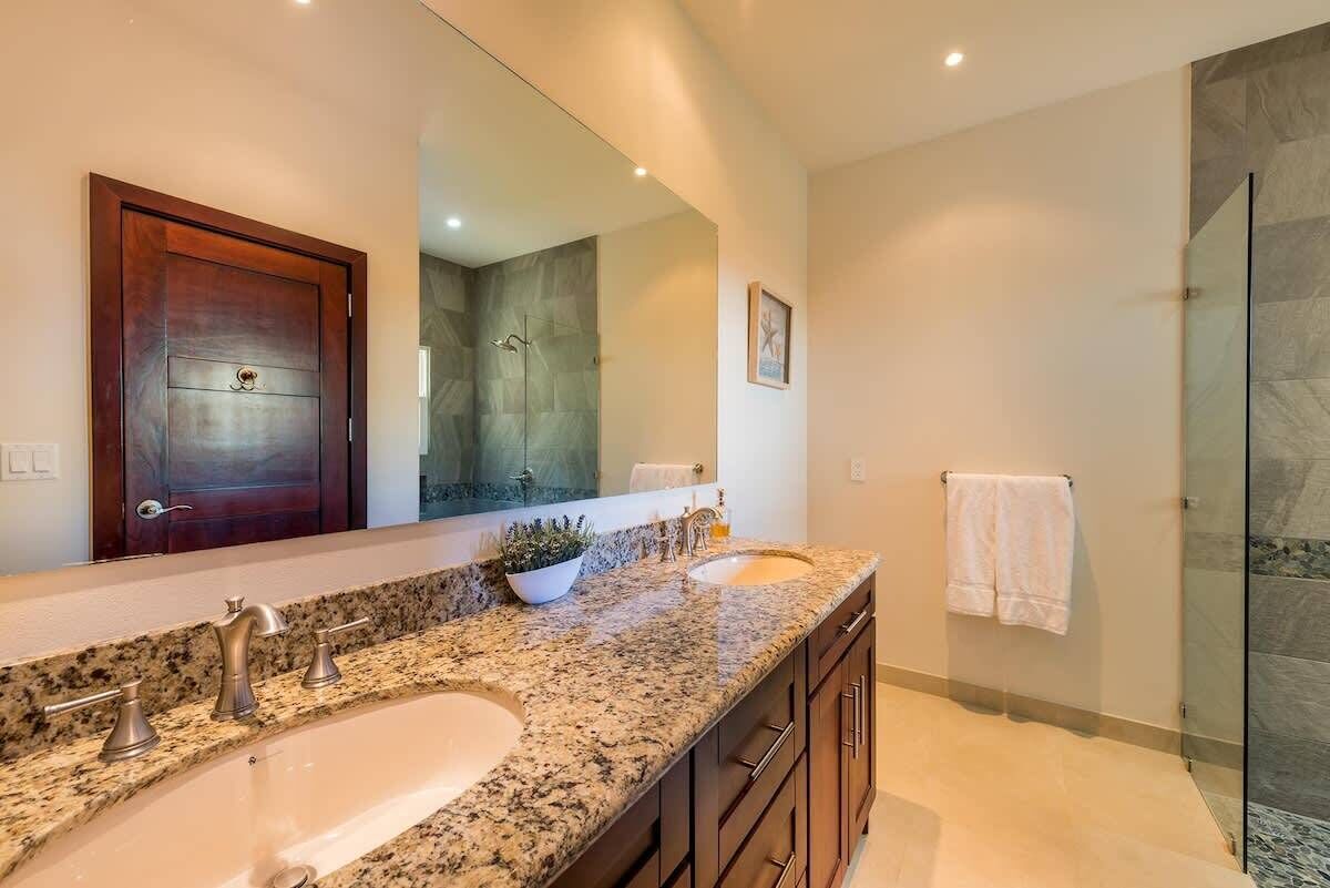 Un baño moderno con encimera de granito, dos lavamanos, un espejo grande y cabina de ducha con paredes de vidrio.
