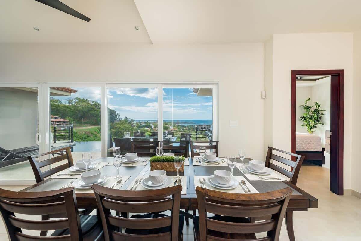 A dining area with a table set for six, large windows offering a view of the landscape, and a door leading to a bedroom.