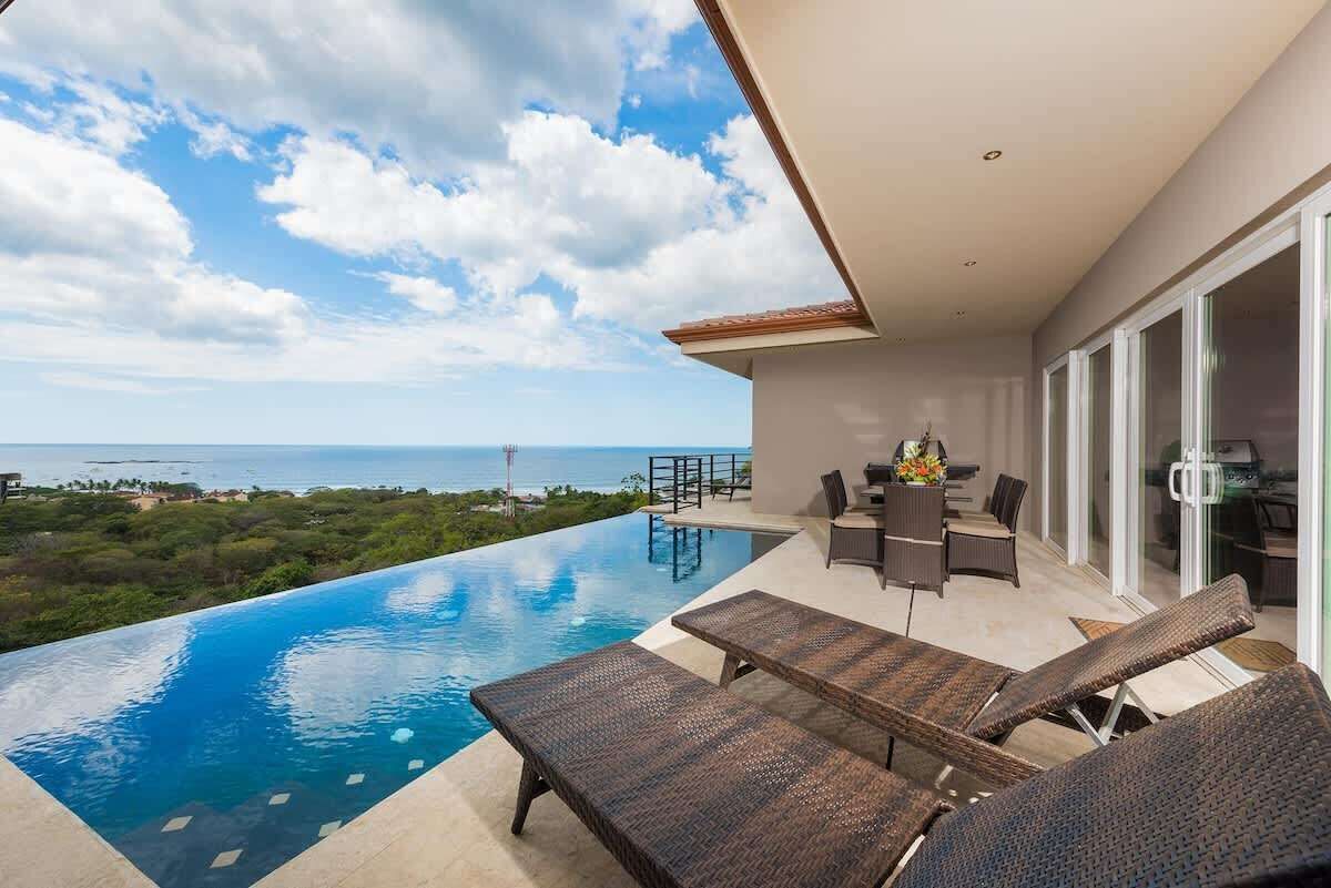 Un área de piscina exterior moderna con sillones de descanso, una mesa de comedor y vistas al océano.
