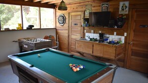 Une table de billard avec des billes, un jeu de fléchettes, un téléviseur et une table de baby-foot dans une pièce aux murs en bois et dotée d’une fenêtre.