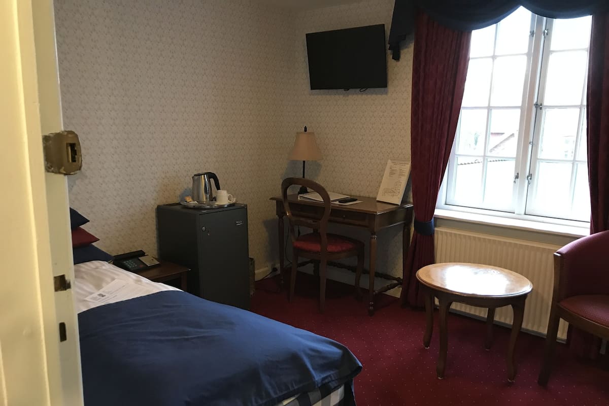 Una habitación de hotel con cama, escritorio con silla, una mesita, televisor en la pared y ventana con cortinas.