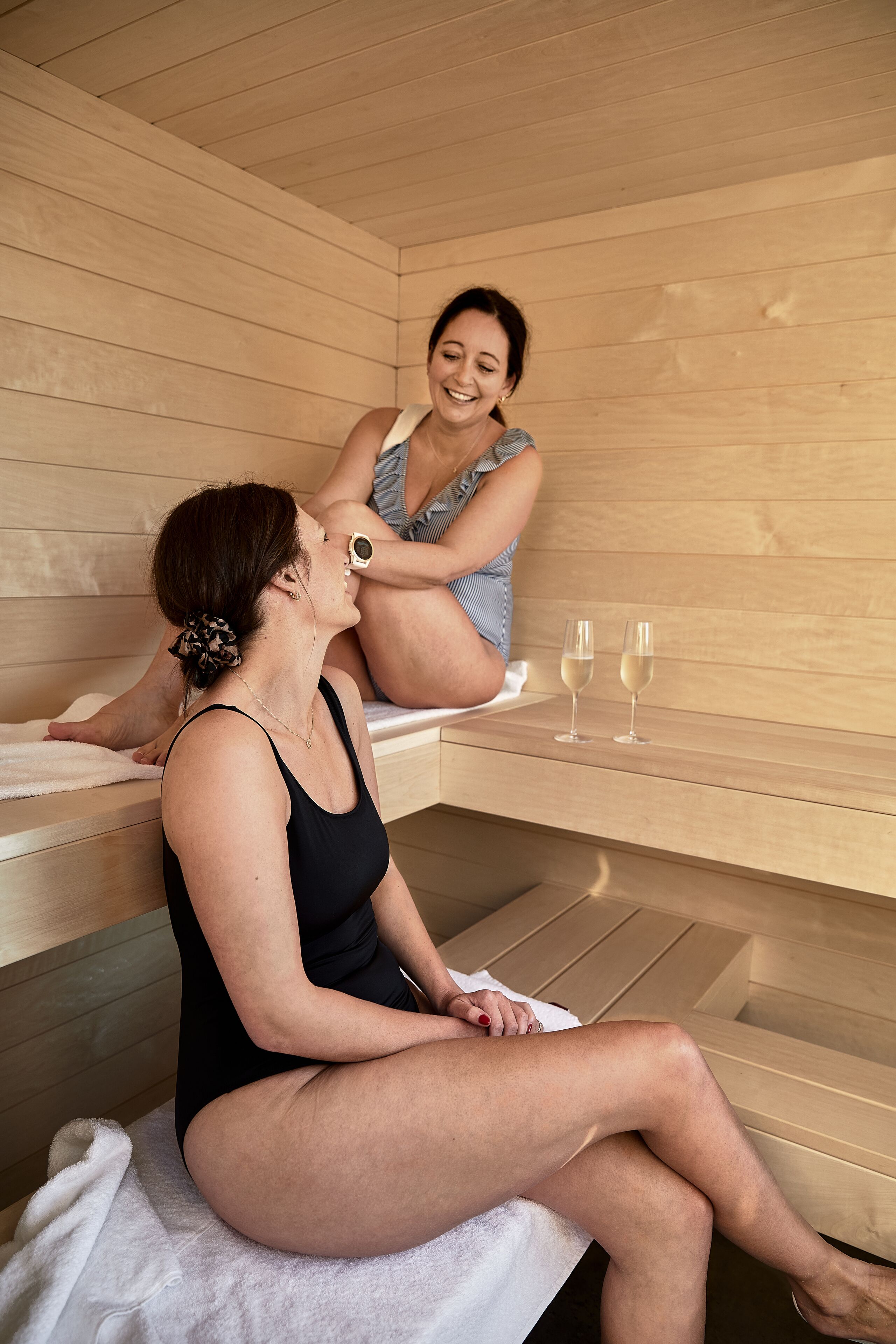 Two people in a sauna with wooden walls and benches, and two glasses of water on the bench.