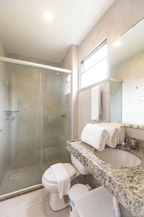 Un baño moderno con ducha envuelta en vidrio, inodoro blanco y lavamanos sobre encimera de granito.