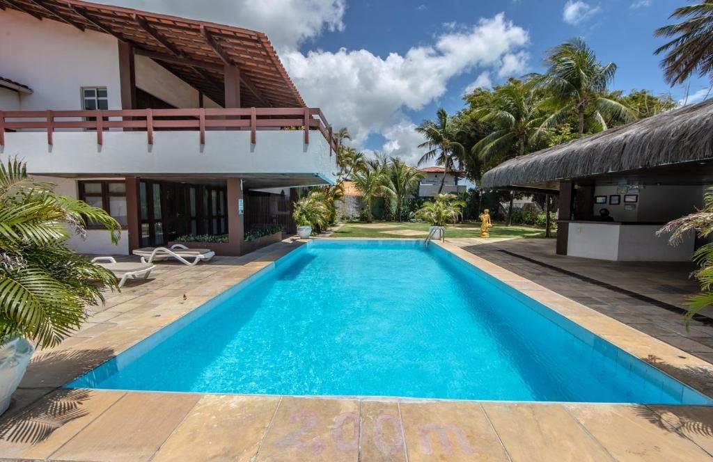 Una piscina con agua cristalina azul, rodeada por un patio con sillones de descanso y un edificio con techo de paja.