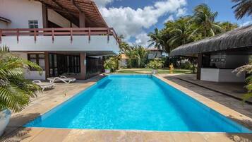 Una piscina con agua cristalina azul, rodeada por un patio con sillones de descanso y un edificio con techo de paja.