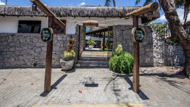 A thatched-roof entrance with wooden posts, a stone wall, and a paved pathway leading to a building with a window and a door. - Duro Beach Garden Hotel (Caucaia)