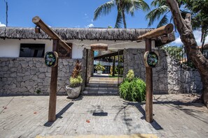 Una entrada con techo de paja, postes de madera, un muro de piedra y un camino pavimentado que lleva a un edificio con una ventana y una puerta.