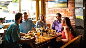 Um grupo de pessoas sentado à mesa de madeira dentro de um restaurante com paredes de tijolos e grandes janelas.