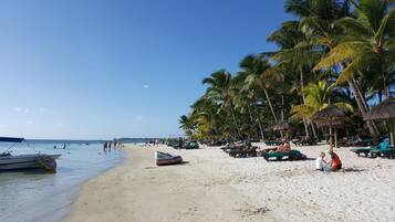 A beach with thatched umbrellas, lounge chairs, and palm trees.