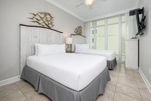 Ein Hotelzimmer mit einem großen Bett, einem kleineren Bett, einem Deckenventilator, einem an der Wand montierten Fernseher und einem Fenster mit Vorhängen.