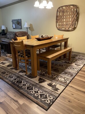A dining area with a wooden table, chairs, and a bench. There is a patterned rug on the floor and a decorative wall piece above the table.