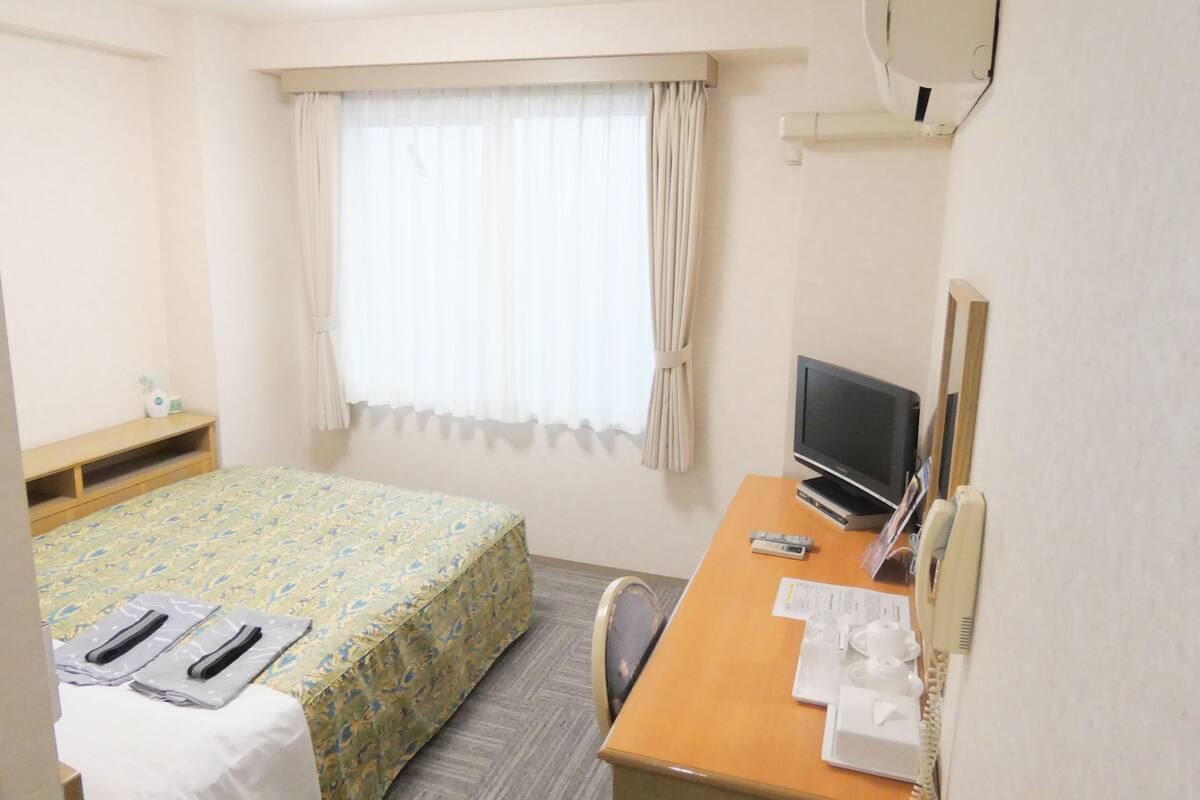 Une chambre d’hôtel avec un lit, un bureau avec un ordinateur, une chaise, une fenêtre avec des rideaux et une petite table sur laquelle se trouvent une bouteille et une télécommande.
