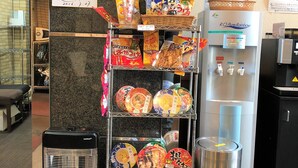 Ein Verkaufsautomatenbereich mit verschiedenen verpackten Snacks und Getränken, einer Kaffeemaschine und einem Mülleimer.