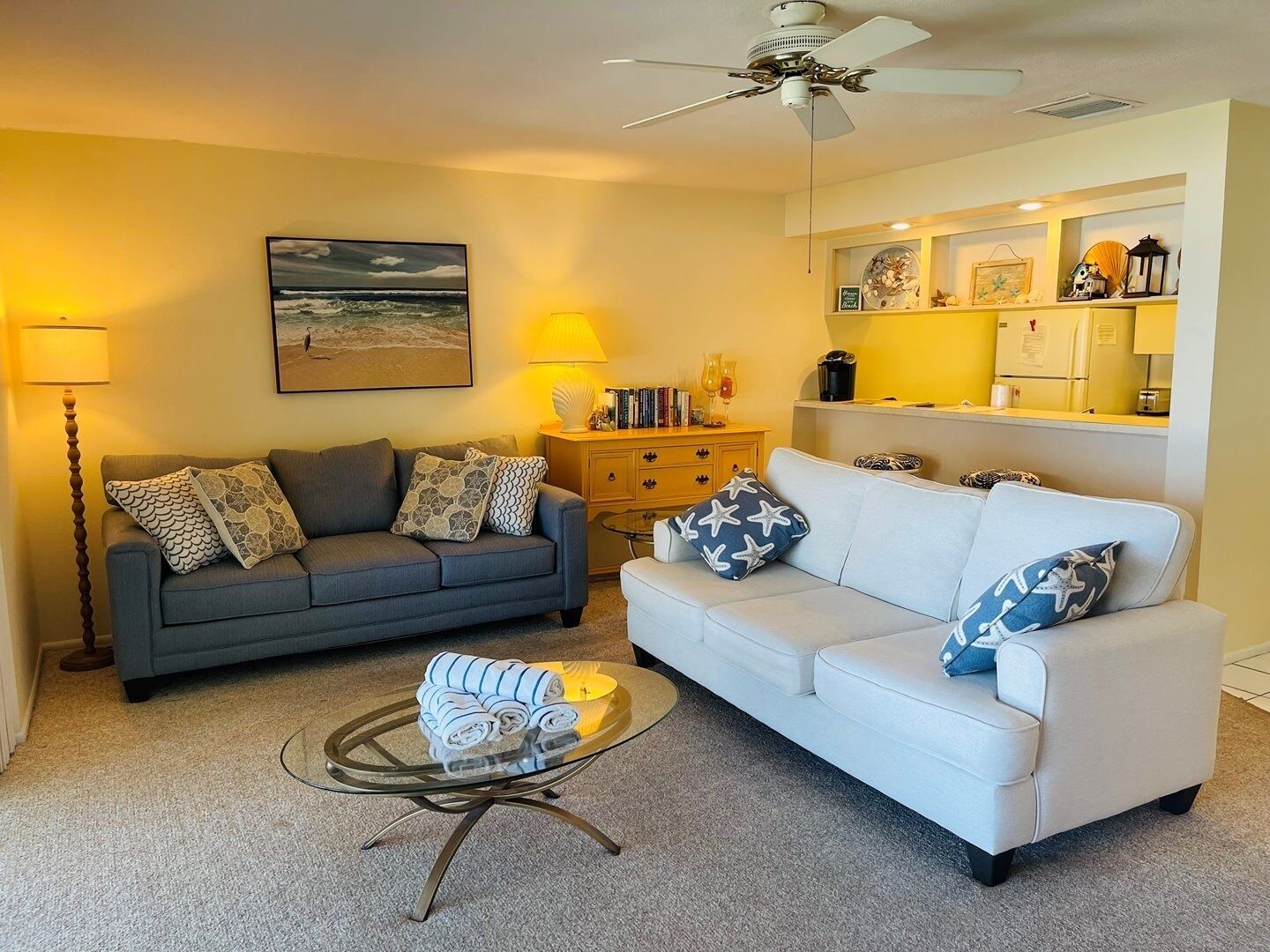 A living room with two sofas, a glass coffee table, a ceiling fan, a painting, and a kitchen area with a refrigerator and shelves.