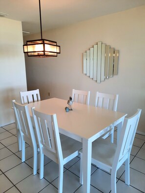 A dining area with a white table and chairs, a hanging light fixture, and a wall-mounted mirror. - Sunset Beach 202 2 Bedroom Condo by RedAwning (Holmes Beach)
