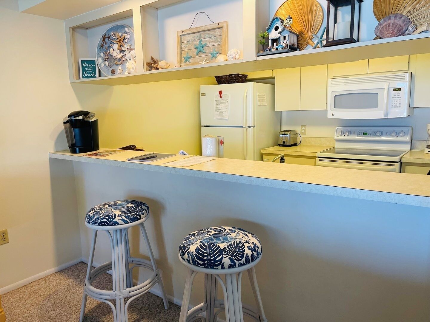 A kitchen with a white countertop, two bar stools, a refrigerator, a microwave, and a toaster.