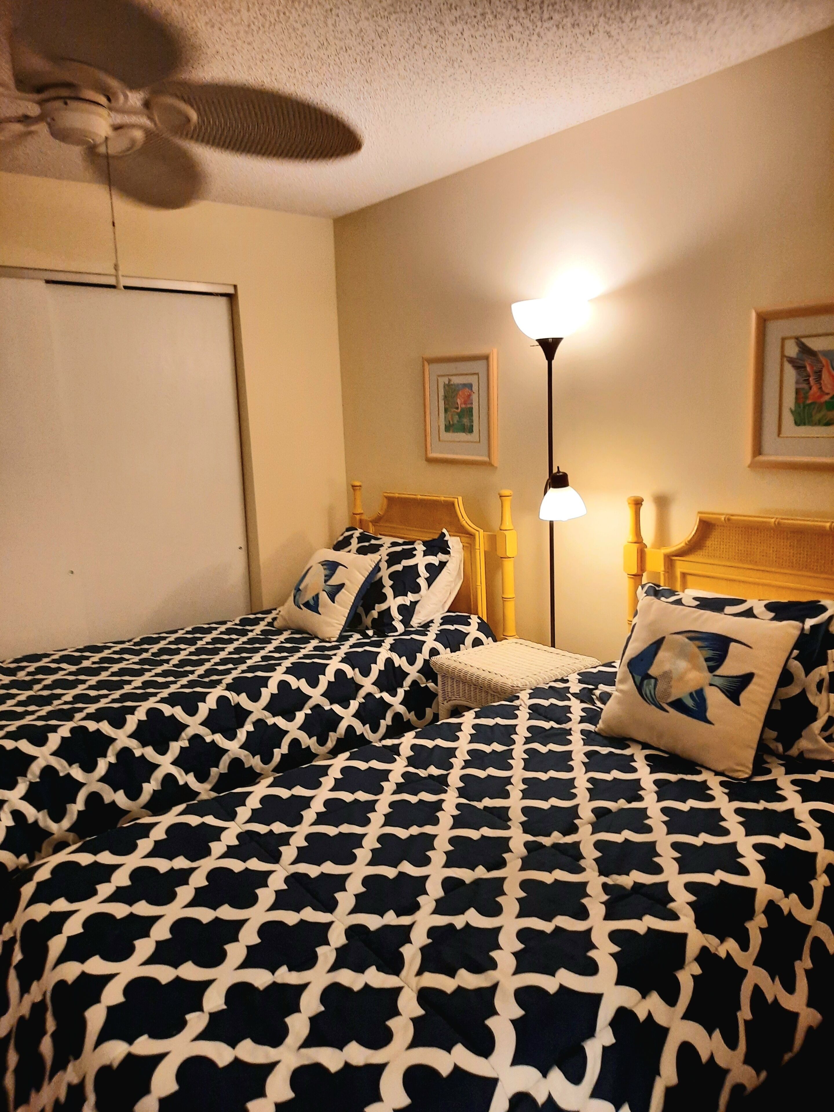 Un dormitorio con dos camas, un ventilador de techo, una lámpara de pie y dos cuadros en la pared.
