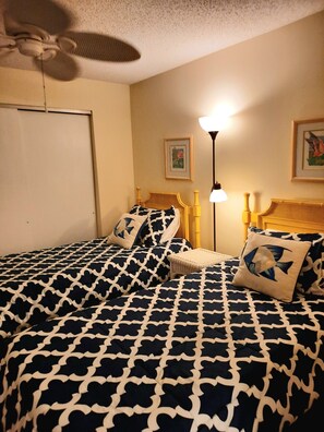 A bedroom with two beds, a ceiling fan, a standing lamp, and two framed pictures on the wall. - Sunset Beach 202 2 Bedroom Condo by RedAwning (Holmes Beach)