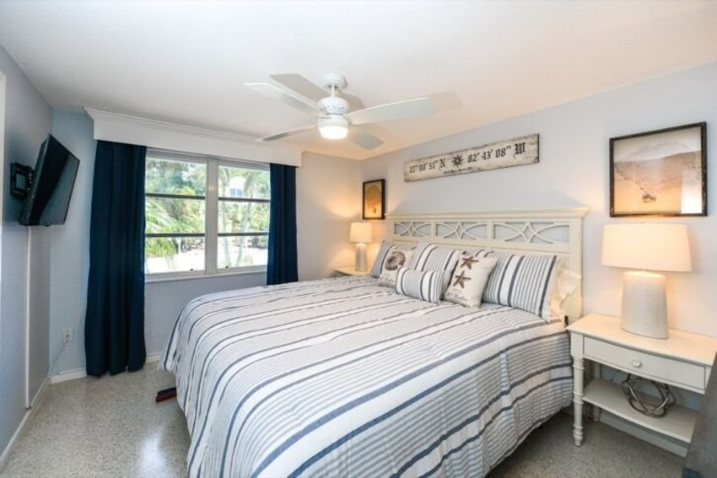 A bedroom with a bed, a ceiling fan, a window with curtains, a bedside table with a lamp, and a wall-mounted TV.
