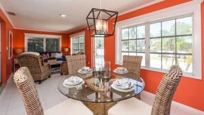 A dining area with a round glass table surrounded by wicker chairs, a hanging light fixture, and large windows with white frames. - 3706 Gulf Dr 2 Bedroom Home by RedAwning (Holmes Beach)