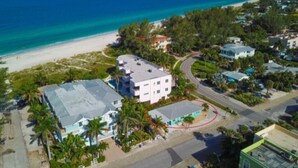 An aerial view of a coastal area with a beach, a large building with a flat roof, and surrounding greenery. - 3706 Gulf Dr 2 Bedroom Home by RedAwning (Holmes Beach)