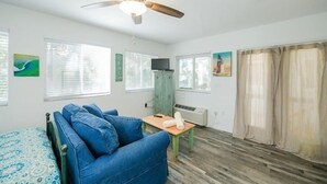 A room with a bed, a blue sofa, a wooden table, a ceiling fan, and a window with curtains. - 3902 Gulf Dr 1 1 Bedroom Apts by RedAwning (Holmes Beach)