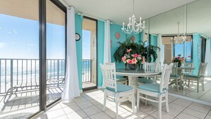 A balcony with a view of the beach, a dining area with white chairs and a table, and a large mirror reflecting the room.