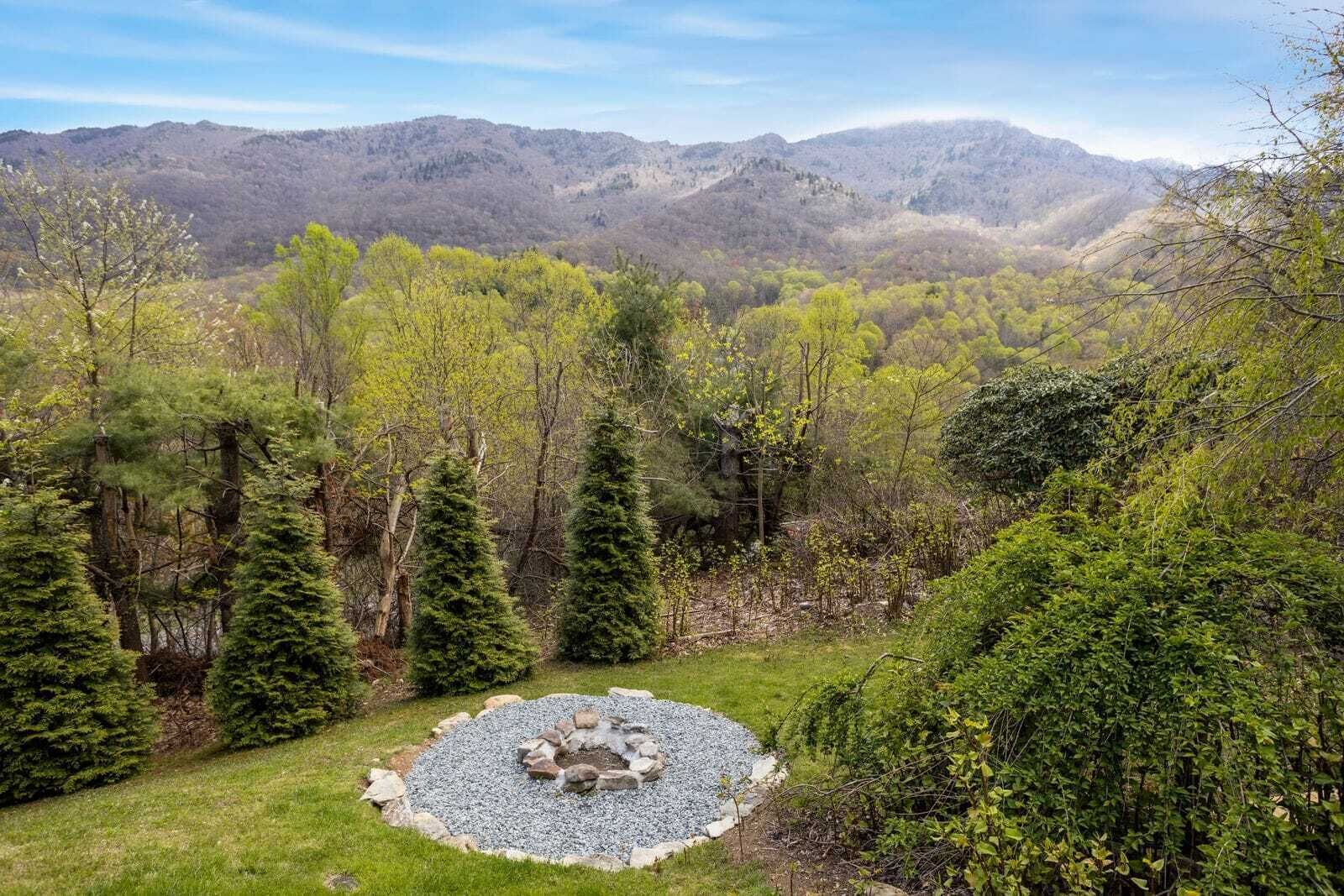 Un foyer circulaire en pierre, entouré d’un cercle de gravier, est aménagé dans une zone herbeuse, bordée d’arbres à feuilles d’aiguilles et dominée par un paysage montagneux.
