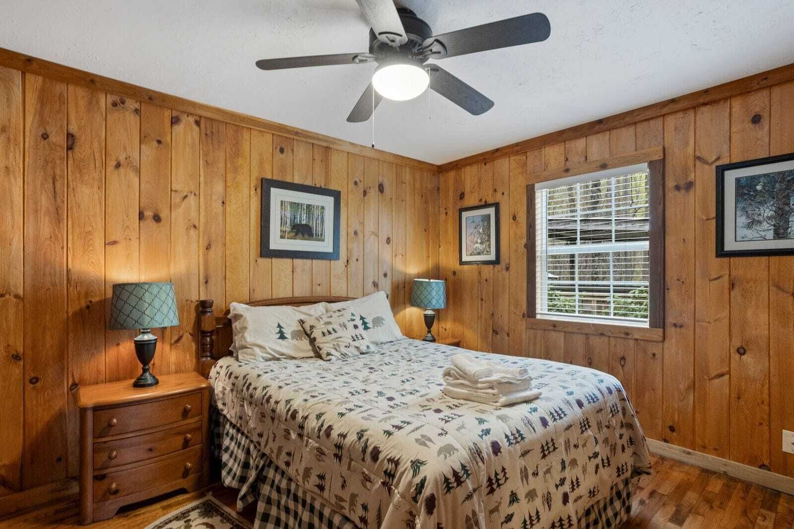 Un dormitorio con paneles de madera, una cama, dos lámparas de noche, un ventilador de techo y una ventana con persianas.