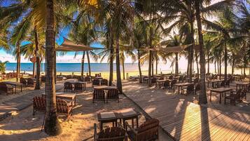 Un restaurante frente a la playa con terraza, palmeras y vista al océano.