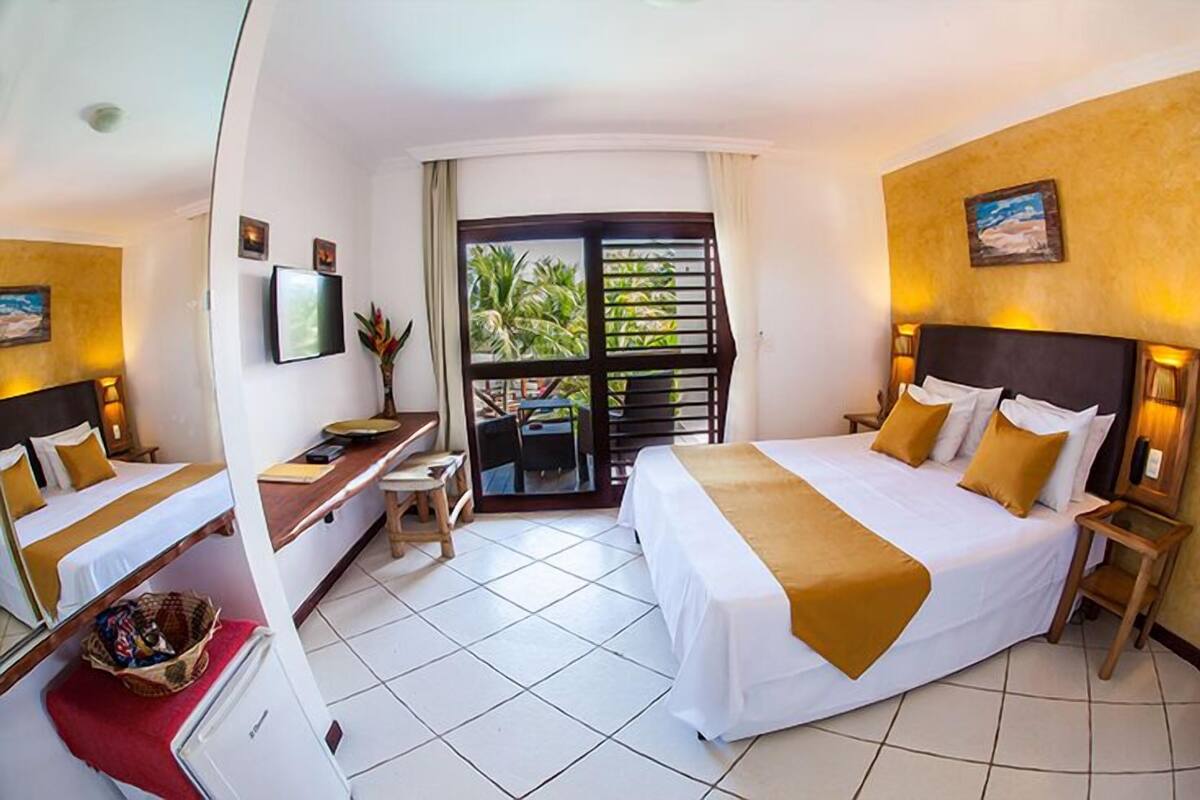 A hotel room with a bed, a desk, a chair, a TV, and a balcony with a view of palm trees.