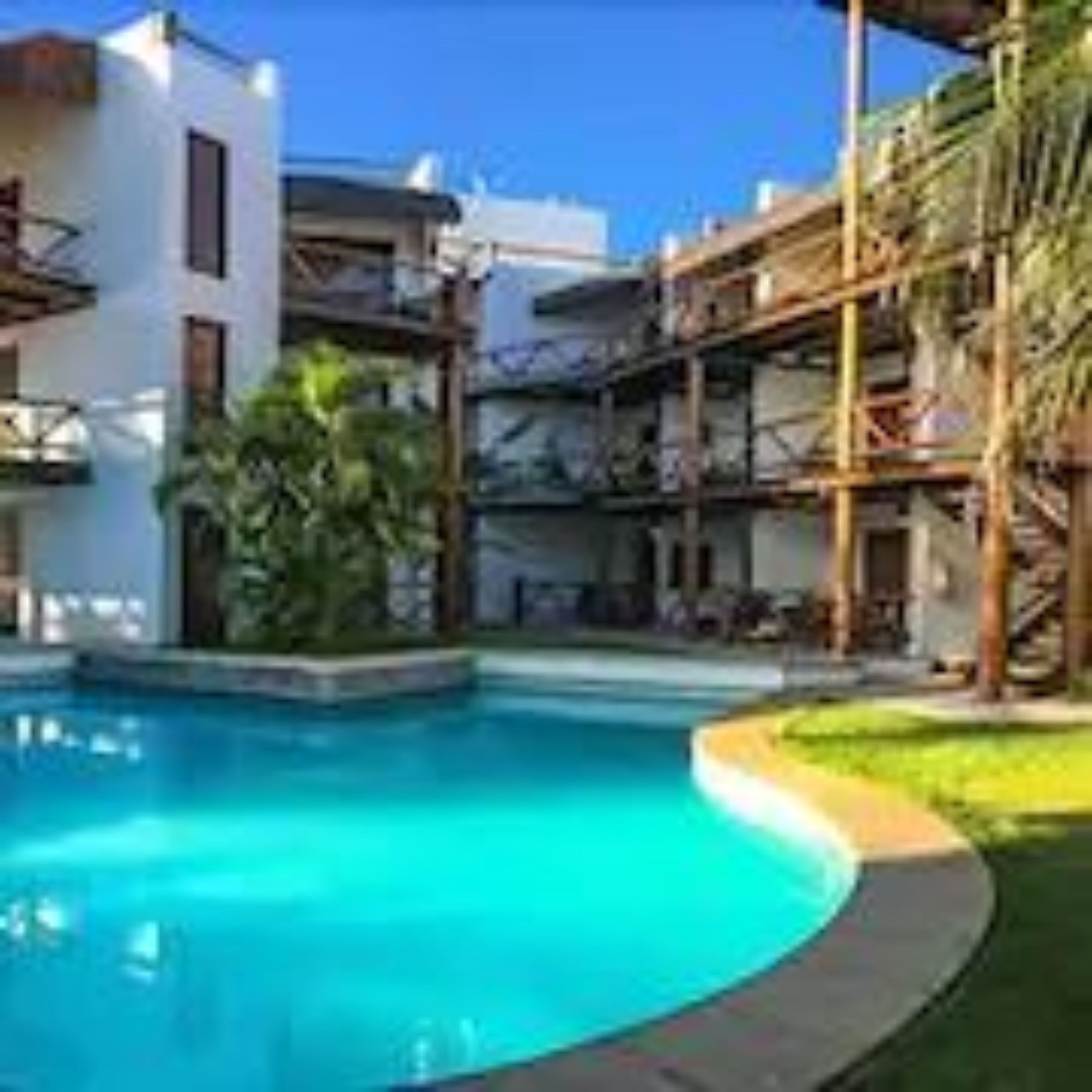 Una piscina con agua cristalina azul, rodeada por una terraza de madera y un edificio blanco con balcones.
