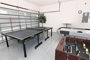 A ping pong table with a TIGA logo, a foosball table, and a plant in a room with white walls and a garage door in the background.