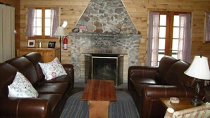 A cozy cabin interior with a stone fireplace, leather sofas, and a ceiling fan.