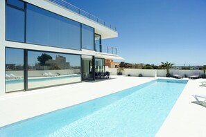 Un área de piscina moderna con una piscina azul cristalina, sillones de descanso y un edificio de dos pisos con amplias ventanas.