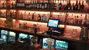 A bar area with shelves of liquor bottles, a cash register, and a refrigerator stocked with beverages. - Lil's Bar and Bistro (York)