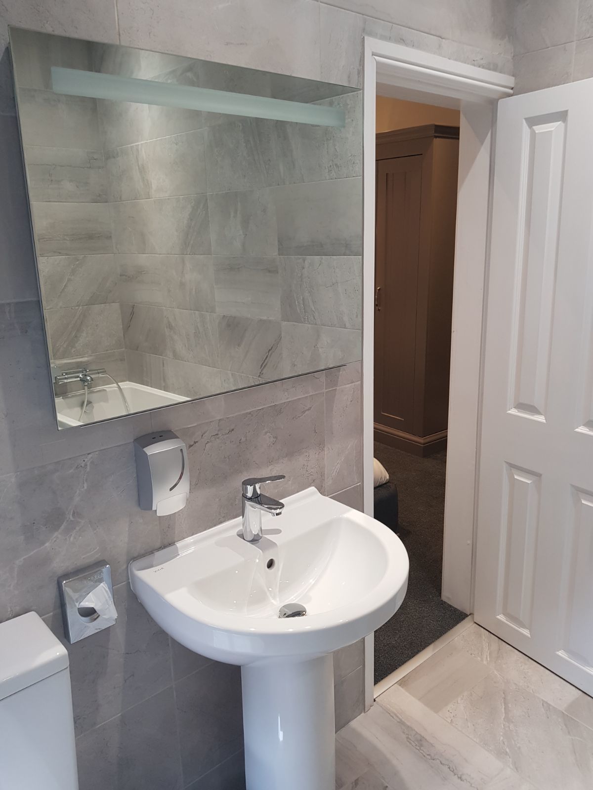 Une salle de bain moderne avec un lavabo sur colonne blanc, une étagère en verre au-dessus du miroir et un mur recouvert de carreaux de marbre.