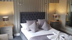 A neatly made bed with a tufted headboard, two bedside tables with lamps, and a large mirror on the wall. - Lil's Bar and Bistro (York)