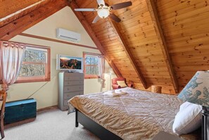 A cozy attic bedroom with a sloped ceiling, a bed with a patterned bedspread, a dresser, a television, and a window with curtains.