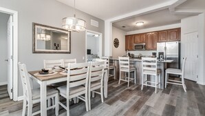 A modern kitchen with wooden cabinets, a stainless steel refrigerator, and a dining area with white chairs and a wooden table. - *BRAND NEW! * CHARMING ORLANDO VISTA CAY  3/BR  2/ FULL BATH CONDO  *SLEEPS 6-8* (Orlando)