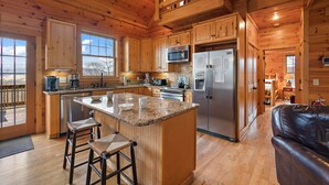 A spacious kitchen with wooden cabinets, granite countertops, and stainless steel appliances. There is a large island with stools and a view of the outdoors through the windows. - Perfect Mountain Getaway with Amazing Views (Banner Elk)