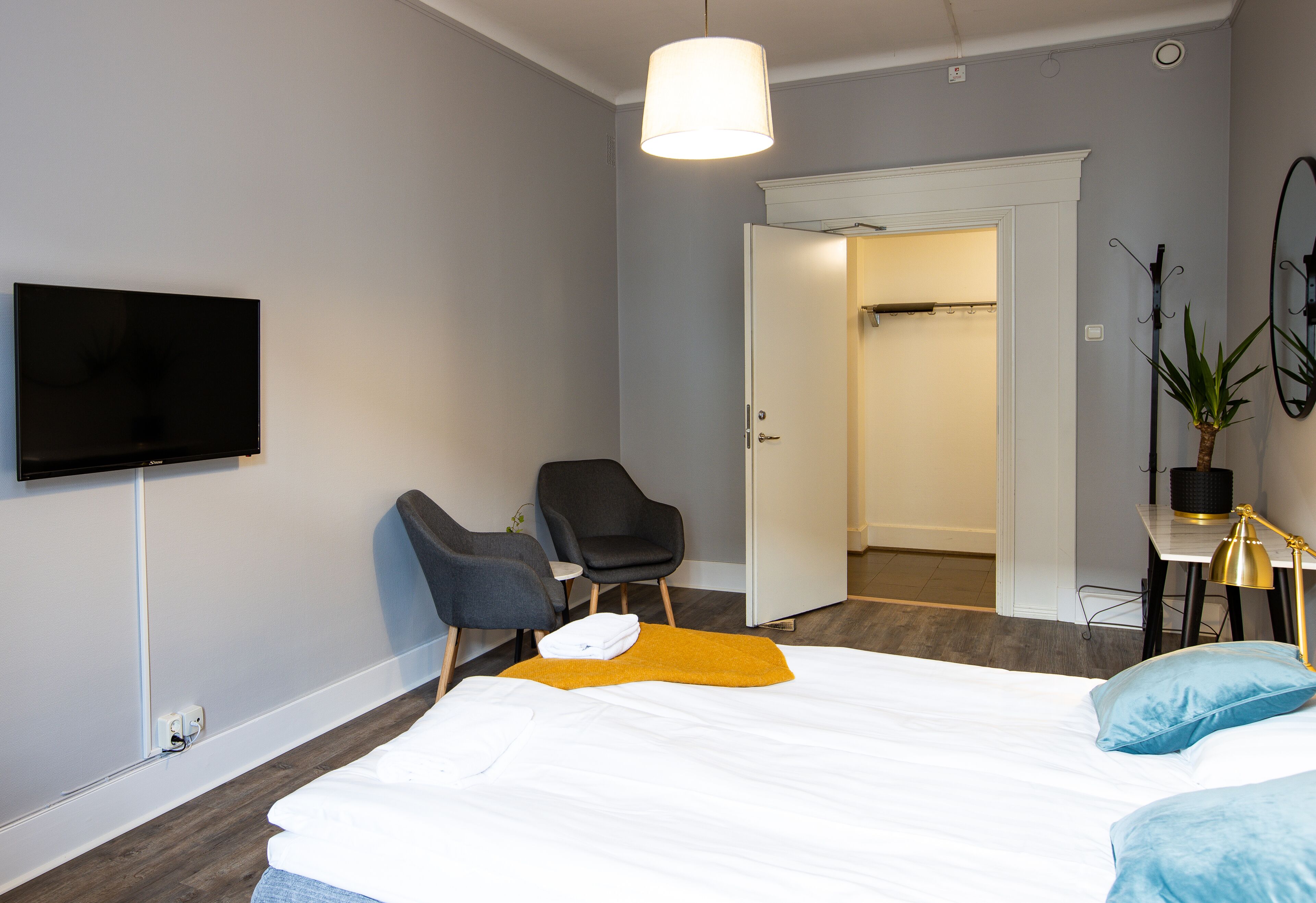 A modern hotel room with a large bed, a flat-screen TV mounted on the wall, a desk with a lamp, and a chair.